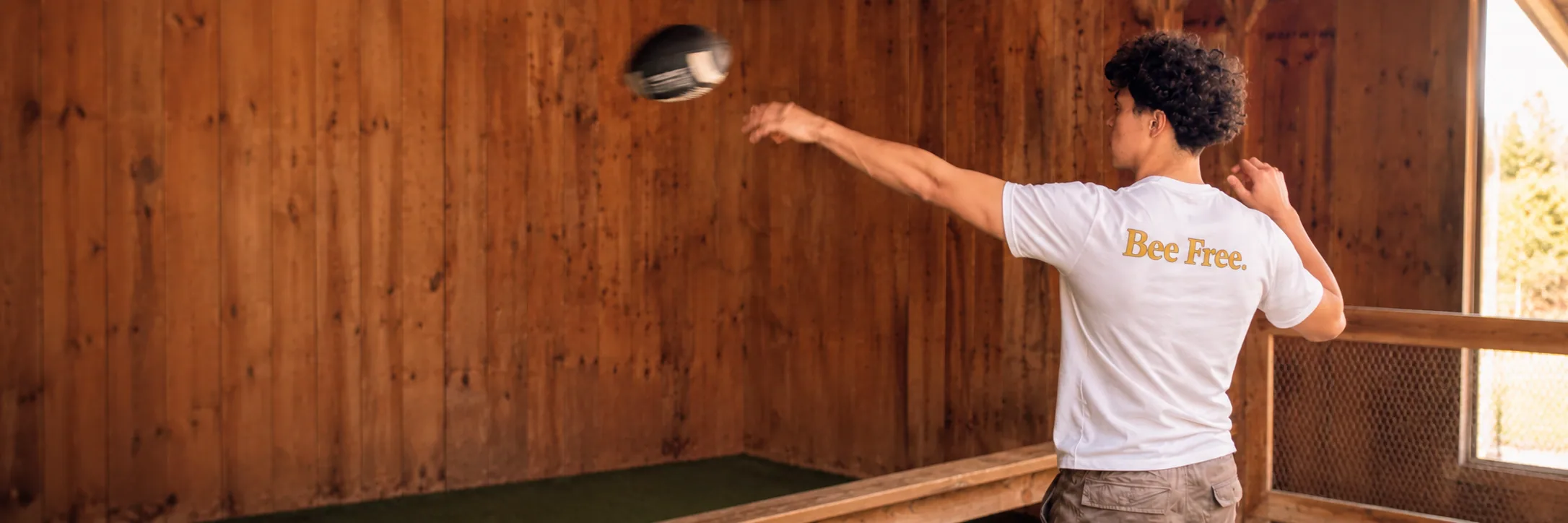 A person in a white Bee Free tee throwing a ball, seen from behind.