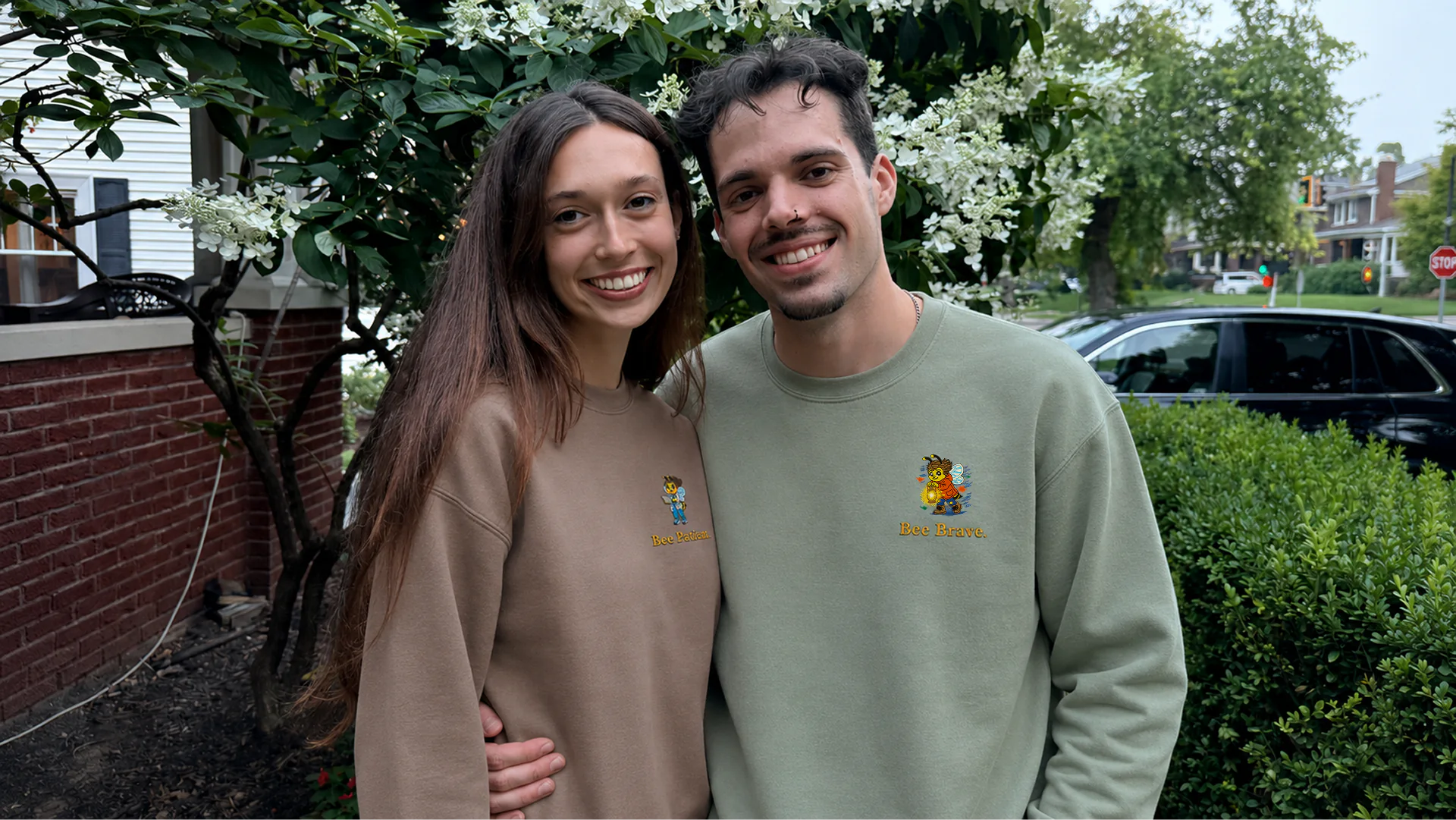 Two adults smiling outdoors in Bee Patient and Bee Brave sweatshirts