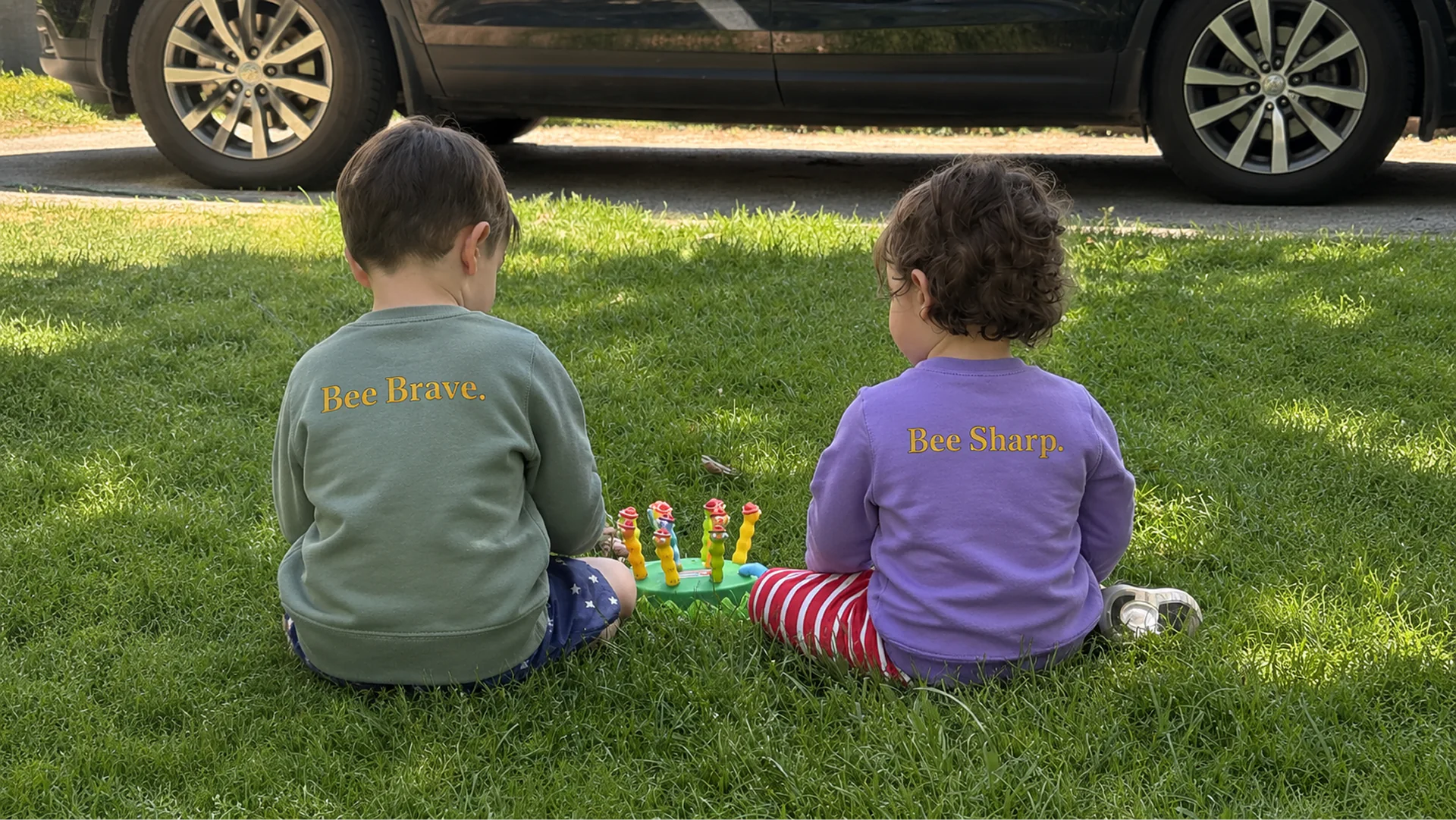 Two children sitting in the grass wearing Bee Brave and Bee Sharp sweatshirts