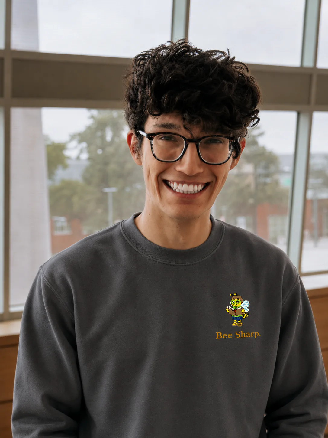 A smiling person wearing a Bee Sharp sweatshirt indoors by large windows